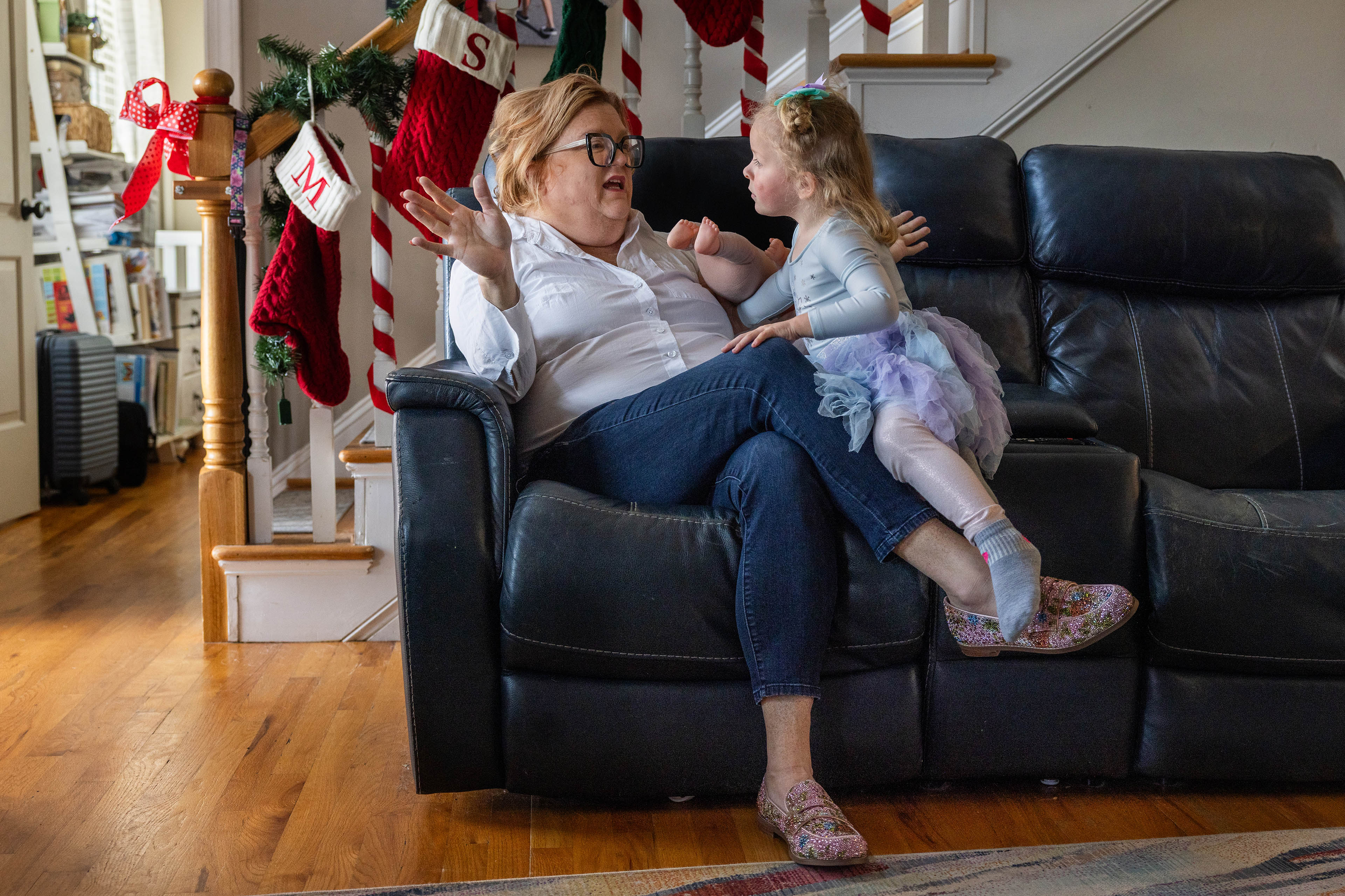 A mother sits with her 3-year-old daughter on a couch in their home. In the background are Christmas decorations.