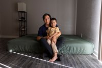 A young boy sits on his father's lap on a mattress covered in a green fitted sheet on the floor.