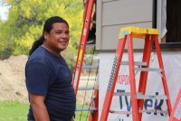 A man with long dark hair pulled into a ponytail and wearing a dark blue t-shirt stands near a building under construction.