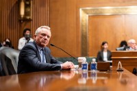 A man in a suit sits at a table in front of a microphone