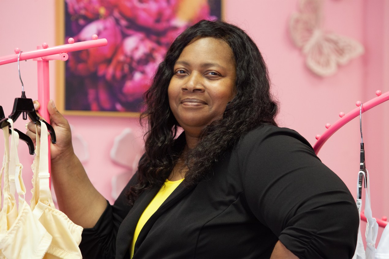 A photo of a Black woman posing for a photo by racks of clothing indoors.