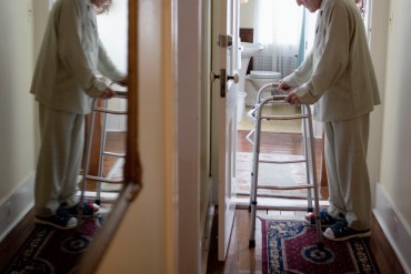 A senior man stands in a hallway with a walker. His reflection is visible, slightly blurred, in a mirror across from him.