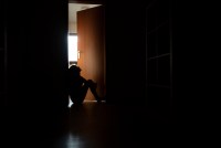 A photo of a teenager sitting in the frame of a doorway backlit by a room filled with daylight.