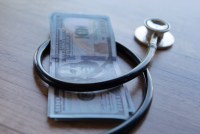 A close-up image of a black stethoscope wrapped around a stack of U.S. dollar bills.