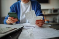 A photo of a person holding receipts and a cell phone while sitting at a table covered in letters and bills.