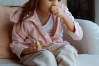 A closely cropped shot of a preschool-aged girl coughing while holding a thermometer.