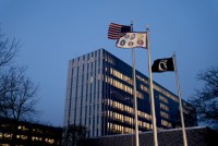 A photo of a large, modern office building in Maryland. In front of it are the American flag, a flag with insignias for six military branches, and the National League of Families POW/MIA flag.