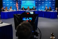 A photo of the CDC's ACIP panel in a room. A television broadcast camera is seen recording the meeting in the center of the frame.