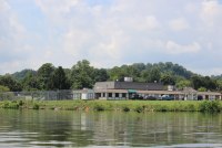 In a photo taken from a craft on a river, a single-story building building sits near the water. There is a fenced-in playground, a shed, a gazebo, and a small parking lot surrounding the building. Trees can be seen in the distance behind the building and its grounds.