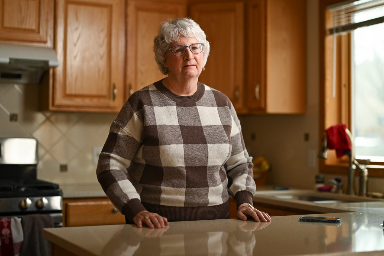 An older woman wearing glasses stands in her kitchen.