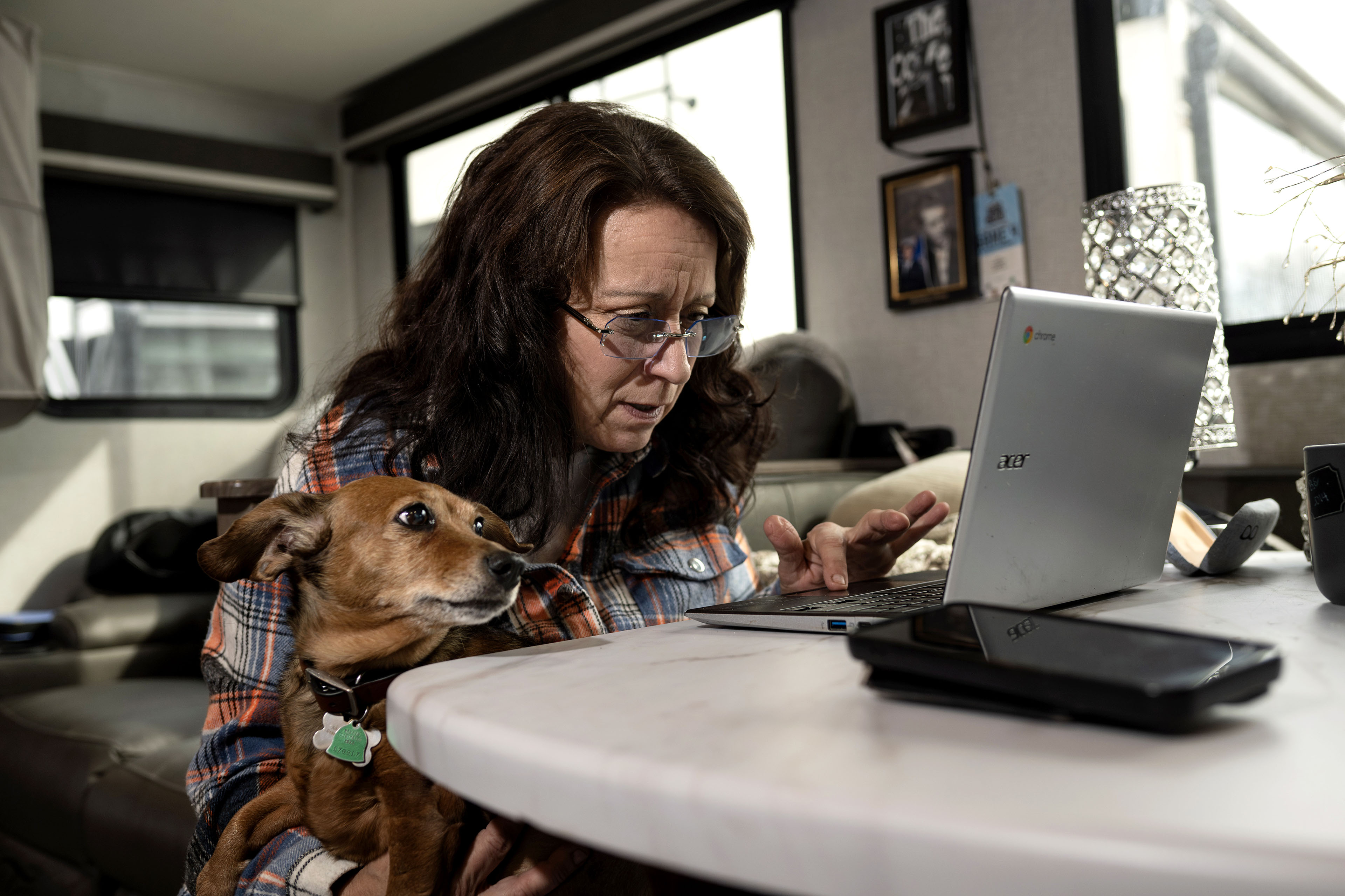 A woman uses a laptop in her kitchen. She wears glasses and leans close to her computer to see. A small dog sits on her lap.