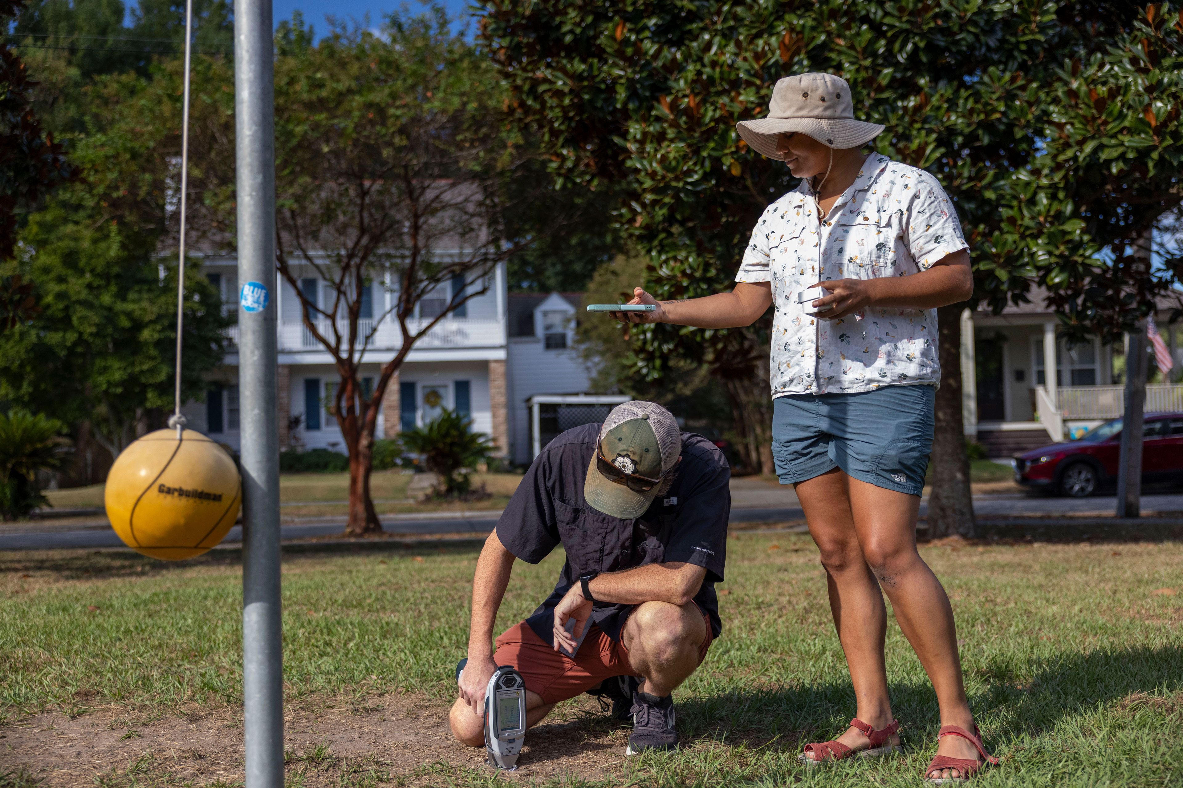 A person squats down to insert a handheld machine into the ground beside a tetherball pole. A second person stands beside the first and holds a phone out over the first person and the location of the sampling.