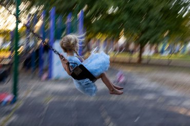 A young child with two braids and a light blue dress with frilly skirt swings on a swing set with barefeet. The rest of the playground and park in the background have a motion blur while the child is in focus.