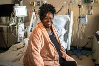 A photo of a woman sitting on a hospital bed with her legs off to the side. She smiles, facing the camera.