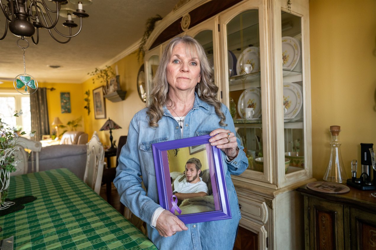 A woman in a light blue denim shirt looks directly at the camera holding a purple picture frame. The photo she is holding shows her son, a young man with a backwards cap, reclining on a couch.