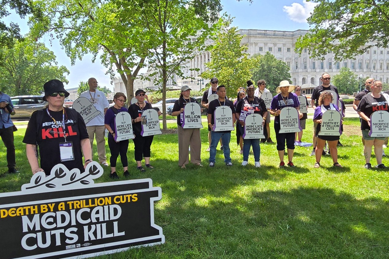 A group of protesters stand holding signs. One large sign reads, "Death by a trillion cuts: Medicaid cuts kill." Others hold signs shaped like tombstones that read, "Here lies America's future," and "R.I.P. Sacrificed for the rich."