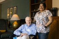 A senior man sits in a chair. A nurse stands beside him with her hand on his back, like good friends. They are both smiling in the direction of the camera.