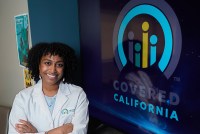 A photo of a woman in a white lab coat posing by a screen with the Covered California logo.