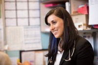 A side portrait of a young woman with straight brown hair. A stethoscope hangs around her neck and she is smiling at something offscreen.