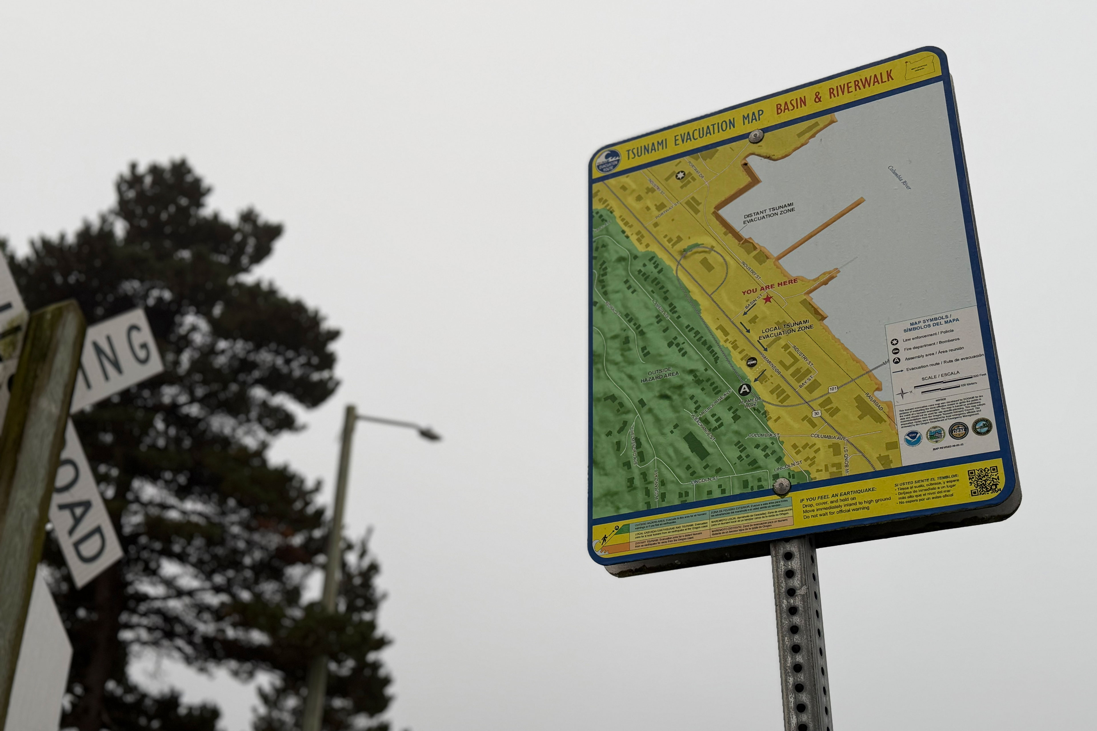 A "Tsunami Evacuation Map" for the Basin & Riverwalk is shown on a metal post. A railroad crossing sign, tree, and streetlight are out of focus behind it.