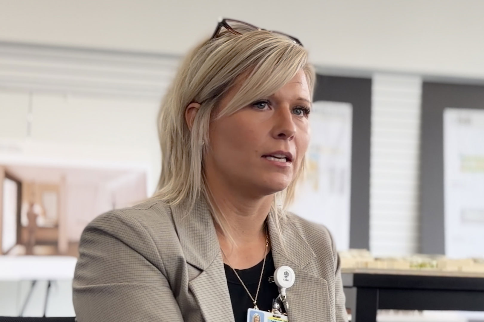 A woman with shoulder-length straight blonde hair sits and speaks to someone just off-camera. She has glasses on top of her head and wears a sandy-colored blazer over a dark shirt.