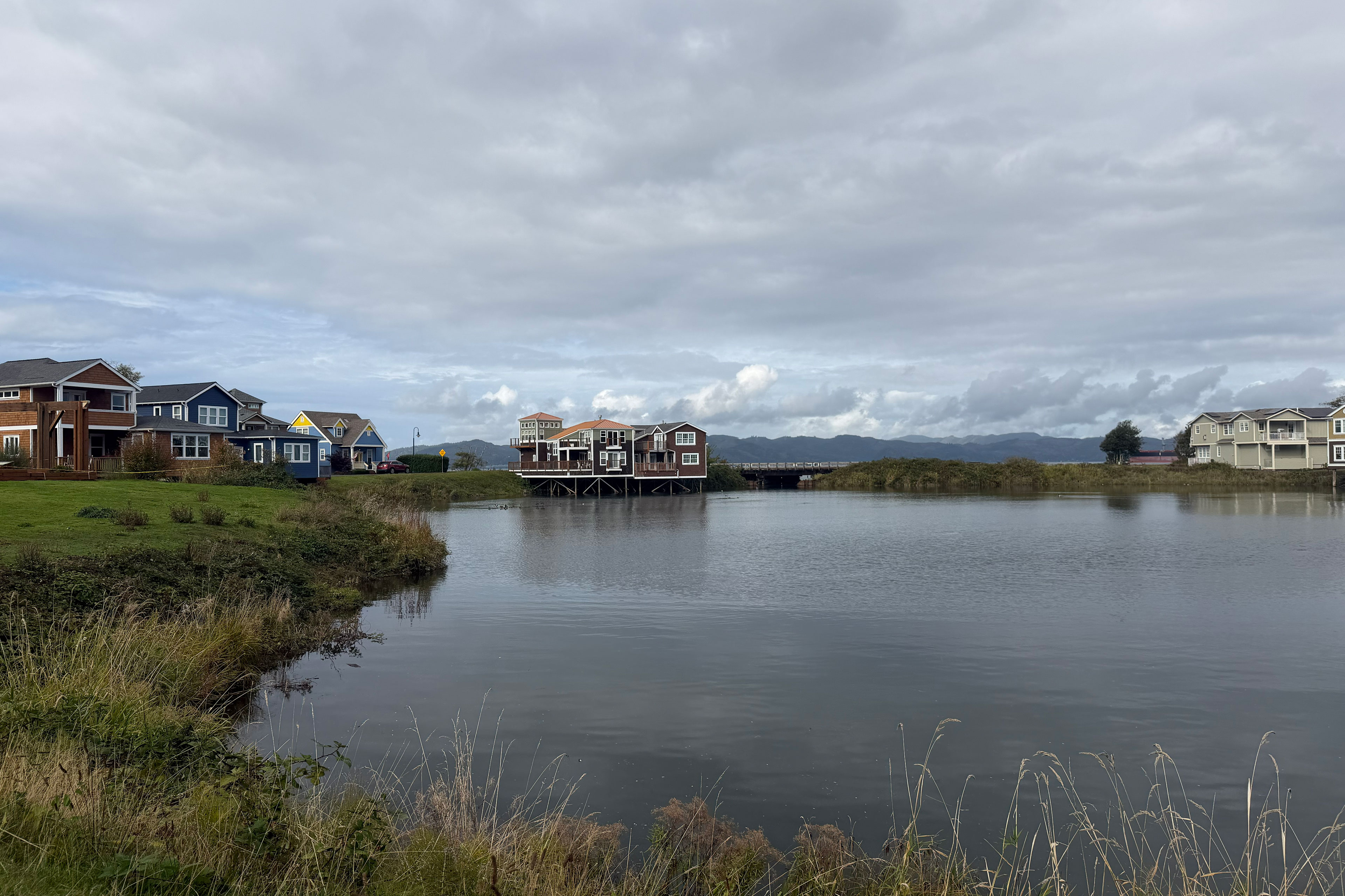 Several houses are show on the waterfront. One in the middle has stilts so that it extends out over the still inlet.
