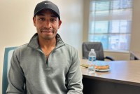 An adult man wearing a baseball cap and a high-collar sweatshirt looks into the camera.